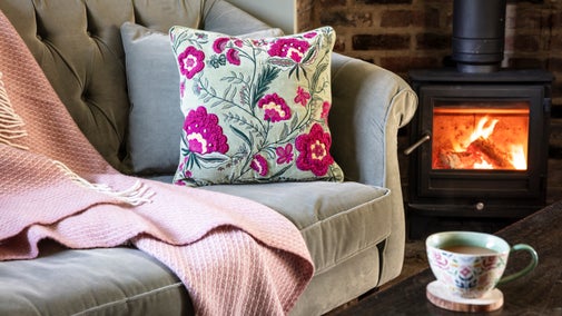 A pink throw and floral cushion displayed on a sage green sofa beside a fireplace from the 2025 autumn/winter collection